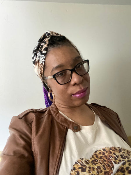 "Multi animal print headband with black, white, and blends of browns, modeled by a moman wearing glasses, gold earrings, leather jacket, off-white t-shirt with animal print lips."
