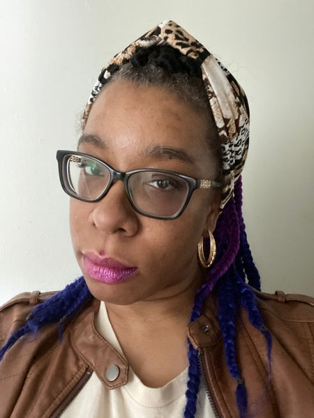 "Multi animal print headband with black, white, and blends of browns, modeled by woman wearing glasses, gold earrings, brown leather jacket, off white shirt "